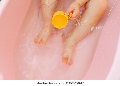 Close Up Unrecognizable Top View Of The Baby Feet In A Pink Baby Bath With Water. The Hands Hold A Toy Yellow Plastic Cup. Copy Space.