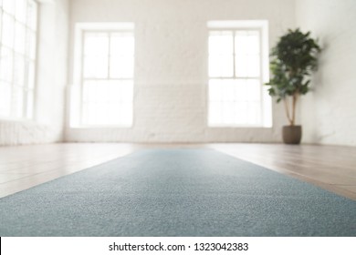 Close up unrolled yoga mat on wooden floor in empty room, modern yoga studio or fitness center with big windows and white brick walls, sport equipment for meditating or exercises