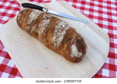 Close Up Of An Unsliced Loaf Of Fresh Homemade Wheat Bread On A Pizza Peel And A Red And White Tablecloth.