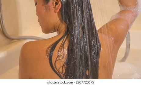 CLOSE UP: Back View Shot Of Beautiful Woman Rinsing Her Hair With Shower Handle. Pretty Young Lady Showering Her Dark Long Hair While Sitting In Bathtub. Time For Personal Hygiene In Home Bathroom.