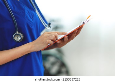 Close Up. A Beautiful Asian Female Doctor Wearing A Blue Nurse Uniform. Hold A Pen And Write Something On The Clipboard To Write Down Patient Information. 