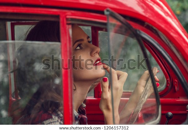 Close up. A beautiful\
pin-up girl in a plaid shirt corrects make-up in the salon of an\
old red retro car.