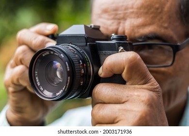 Close up, both hands of an elderly Thai man holding an old film camera and staring to photograph something in a forest garden of rural Thailand.