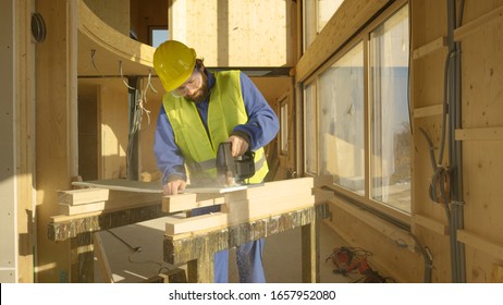 CLOSE UP: Caucasian Worker Building A Beautiful Modern Hardwood House Is Trimming A Gypsum Wallboard. Young Bearded Builder Working In A Cross-laminated House Cuts A Plaster Wall Panel With A Jigsaw.