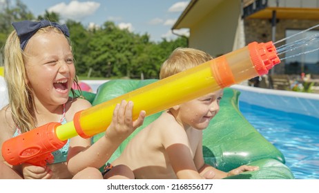 CLOSE UP: Cheerful Brother And Sister In Swimming Pool Spraying With Water Guns. Happy Children Laughing And Enjoying Water Game At Backyard Pool Party. Splashing Water For Refreshment In Summertime.