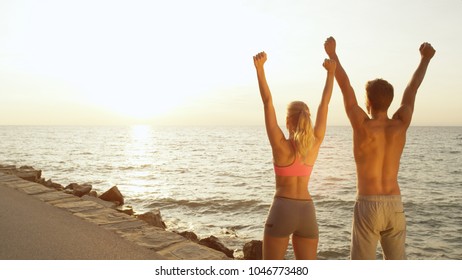 CLOSE UP, COPY SPACE: Young Fit Couple Finishes Scenic Run At The Seaside And Raises Their Arms. Athletic Boy And Girl Celebrate Successful Run Near Rocky Beach On Perfect Day In Summer.