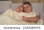 CLOSE UP: Cute couple snuggling under blanket on a couch and watching television. Young pair sitting on sofa and enjoying at relaxing comedy night in comfortable home living room on an autumn evening.
