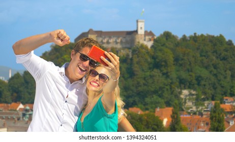 CLOSE UP, DOF: Cheerful Tourist Couple Filming A Vlog From A Rooftop In Beautiful Downtown Of Ljubljana. Happy Man And His Girlfriend Taking Selfies In Front Of The Old Castle On The Green Hill.