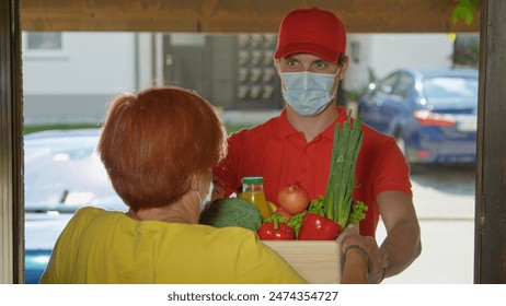 CLOSE UP, DOF: Man wearing a facemask brings fresh organic produce to senior lady. Young courier in a red shirt walks away after delivering groceries to an elderly woman during the covid-19 lockdown. - Powered by Shutterstock