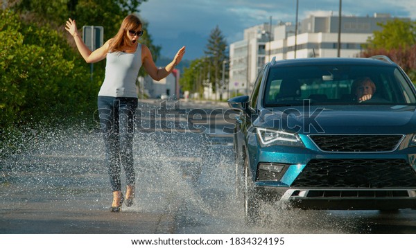 CLOSE UP, DOF: Thoughtless senior driver drives\
her metallic blue car into a puddle, splashing water at the young\
female pedestrian walking along the empty sidewalk in her brand new\
high heels.