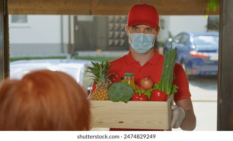 CLOSE UP, DOF: Young delivery guy brings a wooden box of groceries to unrecognizable elderly lady during the coronavirus lockdown. Male courier brings organic produce straight to senior woman's door. - Powered by Shutterstock