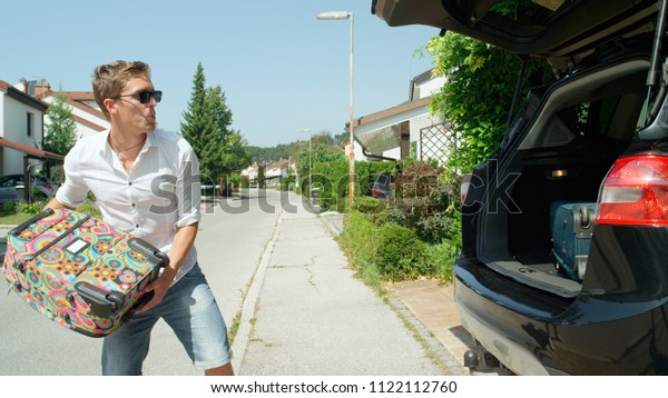 CLOSE UP: Excited young man throws his luggage in\
the back of his big black SUV. Happy traveler living in the sunny\
suburbs throwing travel bags into his car before setting off on a\
fun road trip.