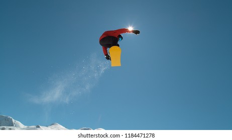 CLOSE UP: Extreme Snowboarder Jumping Across The Clear Blue Sky In Sunny Mountain Wilderness. Freestyle Boarder Snowboarding And Flying Over Camera In Mountain Ski Resort. Snowboard Jump