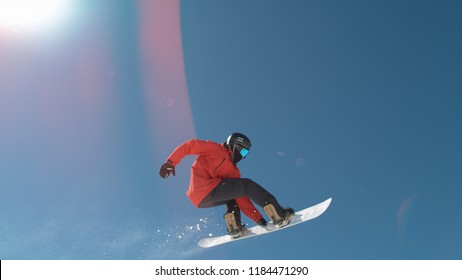 CLOSE UP: Extreme Snowboarder Jumping Across The Clear Blue Sky In Sunny Mountain Wilderness. Freeride Boarder Snowboarding And Flying Over Camera In Mountain Ski Resort. Snowboard Jump