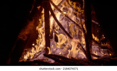 CLOSE UP: Flaming Pieces Of Wood Pilled Up In A Bonfire And Smouldering Embers. High Contrast View Of Burning Logs Against A Black Background. Fiery Flame And Smoke Swirling Around Wooden Remains.