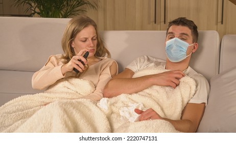 CLOSE UP: Guy With Flu Watching TV And Lady Disinfecting Surroundings With Spray. Couple Covered With Blanket On Comfy Couch And Man Recovering From Flu. Winter Colds And Viruses Spreading Around.