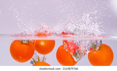 CLOSE UP, HALF UNDERWATER: Cinematic Shot Of Small Red Tomatoes Falling And Splashing Into The Cold Fresh Water. Vibrant Ripe Cherry Tomatoes Fall Into The Crystal Clear Fresh Water. Washing Produce.