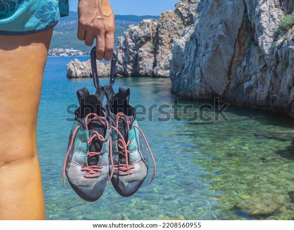 CLOSE UP: Held climbing shoes with climbing\
boulder above seawater in background. Young woman holding sports\
shoes tied with a carabiner before going deep water solo climbing\
at the beautiful seaside.