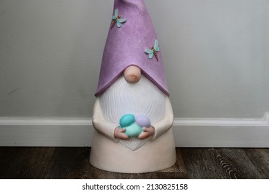 The close up, isolated image of a garden gnome wearing a purple hat that is covered in dragonflies. The decoration has a beard and is holding colorful Easter eggs.