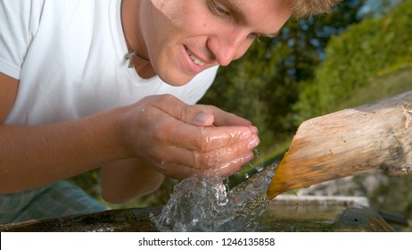 CLOSE UP: Joyful Caucasian Male Tourist Freshening Up By Washing His Face During His Hike With Crystal Clear Stream Water Coming Out Of The Wooden Pipe. Man Lifts Up A Handful Of Pure Water To Drink.