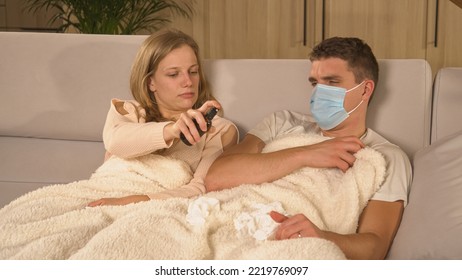 CLOSE UP: Lady Disinfecting Surroundings With Spray And Guy With Flu Watching TV. Couple Covered With Blanket On Comfy Couch And Man Recovering From Flu. Winter Colds And Viruses Spreading Around.