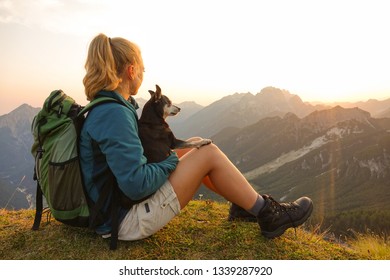 CLOSE UP, LENS FLARE: Carefree blonde haired girl sits on the grassy mountaintop and pets her dog while observing the picturesque nature at sunset. Happy woman rests with puppy after a morning hike.