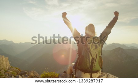 CLOSE UP, LENS FLARE: Ecstatic trekker girl celebrates reaching the mountaintop with a breathtaking view of the mountain range on a sunny summer day. Unrecognizable hiker woman outstretches arms.