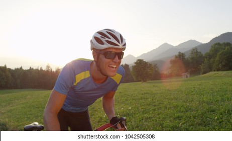 CLOSE UP, LENS FLARE: Exhausted Professional Male Cyclist Pushing Himself To The Limit As He Races Across The Countryside On His Road Bike At Golden Sunset. Cycling Athlete Pedaling Up A Sunny Road.