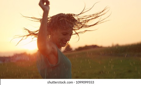 CLOSE UP, LENS FLARE: Joyful blonde girl enjoys her evening in the countryside by dancing in the rain. Stunning golden sun rays shine on playful young woman spinning and enjoying a spring shower.