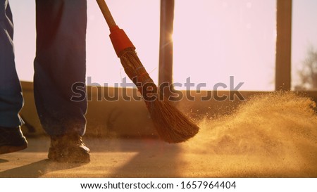 CLOSE UP, LENS FLARE, LOW ANGLE: Contractor is sweeping the dirty floor with a retro straw broom at scenic golden sunset. Worker sweeps the dusty floor after a long day at a busy construction site.