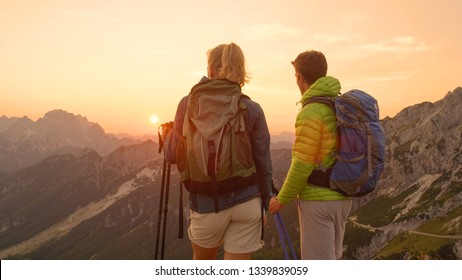 CLOSE UP, LENS FLARE: Unrecognizable Hiker Couple Watching The Sunset From The Scenic Mountaintop In Julian Alps. Cheerful Man And Woman Observe The Golden Sunrise During Their Fun Summer Hiking Trip.