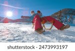 CLOSE UP, LOW ANGLE, LENS FLARE: Happy Caucasian woman speeds down the snowy hill on a wooden sled. Sparkling snowflakes fly into the camera as smiling young female tourist sleighs down a steep hill.