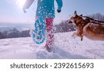 CLOSE UP, LOW ANGLE VIEW: Lady and her playful dog have fun running in snow. Woman is running with snowshoes with her brown doggo by side. Beautiful sunny winter day for outdoor sports activities.