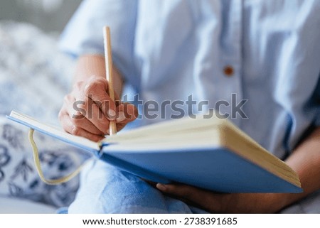 close up, middle aged White woman hand holding pencil, writing in blue notebook on bed, soft natural light, cozy home bedroom, mindful journaling, personal goals, quiet self care moment.