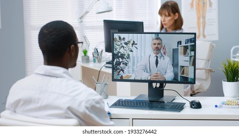 Close Up. Over Shoulder View Of African American Man Doctor Sitting In Cabinet In Clinic Speaking With Caucasian Young Coworker Consulting Online On Computer, Medical Web Conference Healthcare Concept