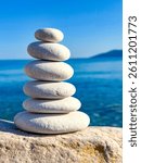 CLOSE UP: Perfectly balanced stack of six smooth white stones is positioned on a rock surface overlooking blue ocean. Wavy sea and clear sky create a tranquil backdrop, evoking peace and mindfulness.