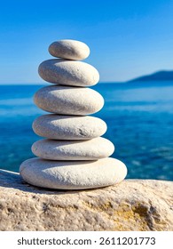 CLOSE UP: Perfectly balanced stack of six smooth white stones is positioned on a rock surface overlooking blue ocean. Wavy sea and clear sky create a tranquil backdrop, evoking peace and mindfulness.