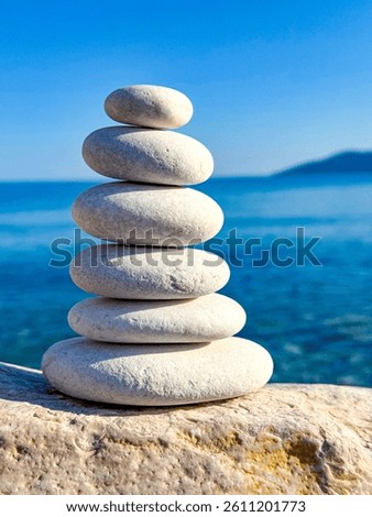 CLOSE UP: Perfectly balanced stack of six smooth white stones is positioned on a rock surface overlooking blue ocean. Wavy sea and clear sky create a tranquil backdrop, evoking peace and mindfulness.