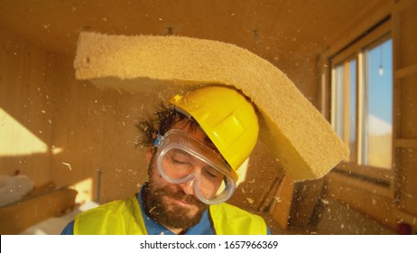 CLOSE UP, PORTRAIT, DOF: Funny Shot Of A Smiling Builder Getting Hit With A Thick Piece Of Foam. Bearded Male Surveyor Wearing A Yellow Helmet Gets Struck In The Head By A Piece Of Falling Insulation.