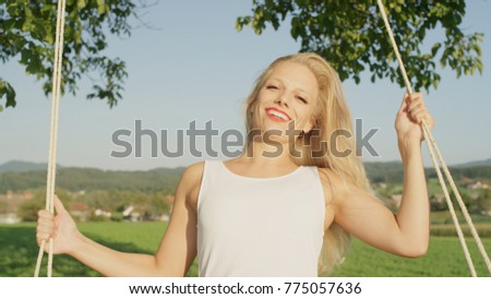 CLOSE UP, PORTRAIT: Smiling woman swaying on a rope swing in nature by herself. Lonely sun kissed blonde girl smiling at camera as she swings away on swing. Young woman glowing in warm summer sunshine