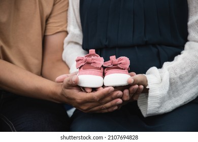 Close up, Pregnant women sit on bed with handsome husband and hold couple red baby shoes on the belly. A gift for the baby to be born in the future.