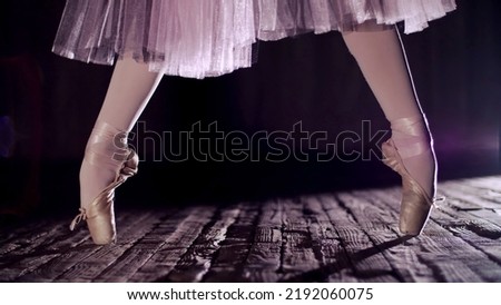 close up, in rays of spotlight, on the stage of the old theater hall. ballerina in white ballet skirt, raises on toes in pointe shoes, performs elegantly a certain ballet exercise, relleve. High