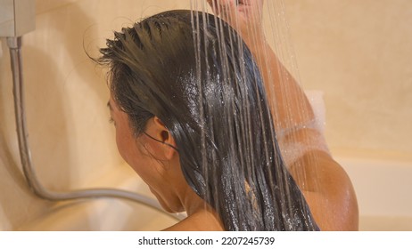 CLOSE UP: Rear View Shot Of Pretty Young Lady Washing Her Hair With Shower Handle Beautiful Woman Showering Her Long Dark Hair While Sitting In Bubble Bath. Time For Personal Hygiene In Home Bathroom.