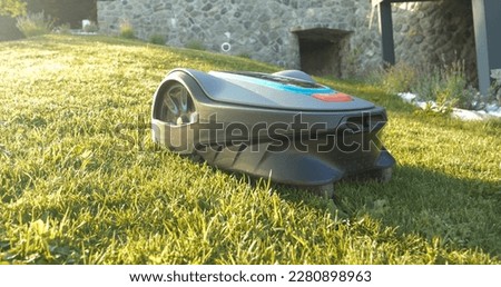 CLOSE UP: Side view of robotic lawn mower trimming green grass in modern garden. Futuristic gardening equipment at work. Lawn robot, illuminated by golden sunlight, cutting green turf in the backyard.