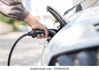 Close up, side view shot, selective focus of a woman's hand grabbing, plugging, holding the type2 EV charger plug handle in front of an electric car hood. Renewable sustainable energy concept.