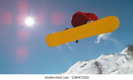 CLOSE UP: Snowboarder Jumping Big Air Kicker, Spraying Snowflakes And Flying Over Sun On Perfect Winter Day. Snowboard Jump In Snow Park. Sunbeams Shining Past Jumping Boarder In Mountains