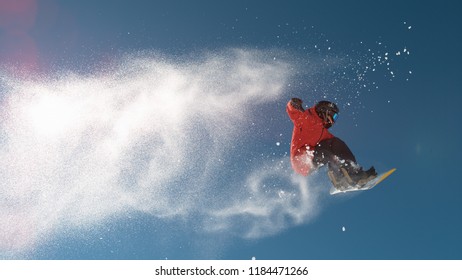 CLOSE UP: Snowboarder Jumping Big Air Kicker, Spraying Snowflakes And Flying Over Sun On Perfect Winter Day. Snowboard Jump In Snow Park. Sunbeams Shining Past Jumping Boarder In Mountains