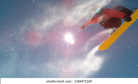 CLOSE UP: Snowboarder Jumping Big Air Kicker, Spraying Snowflakes And Flying Over Sun On Perfect Winter Day. Snowboard Jump In Snow Park. Sunbeams Shining Past Jumping Boarder In Mountains