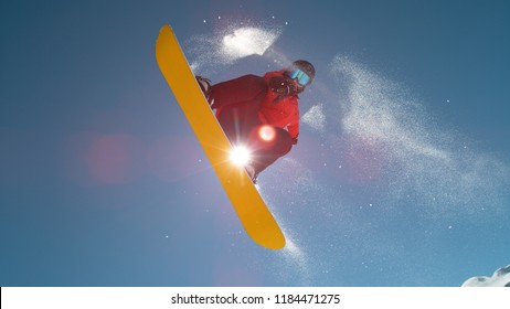 CLOSE UP: Snowboarder Jumping Big Air Kicker, Spraying Snowflakes And Flying Over Sun On Perfect Winter Day. Snowboard Jump In Snow Park. Sunbeams Shining Past Jumping Boarder In Mountains