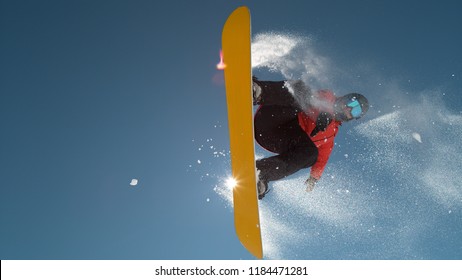 CLOSE UP: Snowboarder Jumping Big Air Kicker, Spraying Snowflakes And Flying Over Sun On Perfect Winter Day. Snowboard Jump In Snow Park. Sunbeams Shining Past Jumping Boarder In Mountains